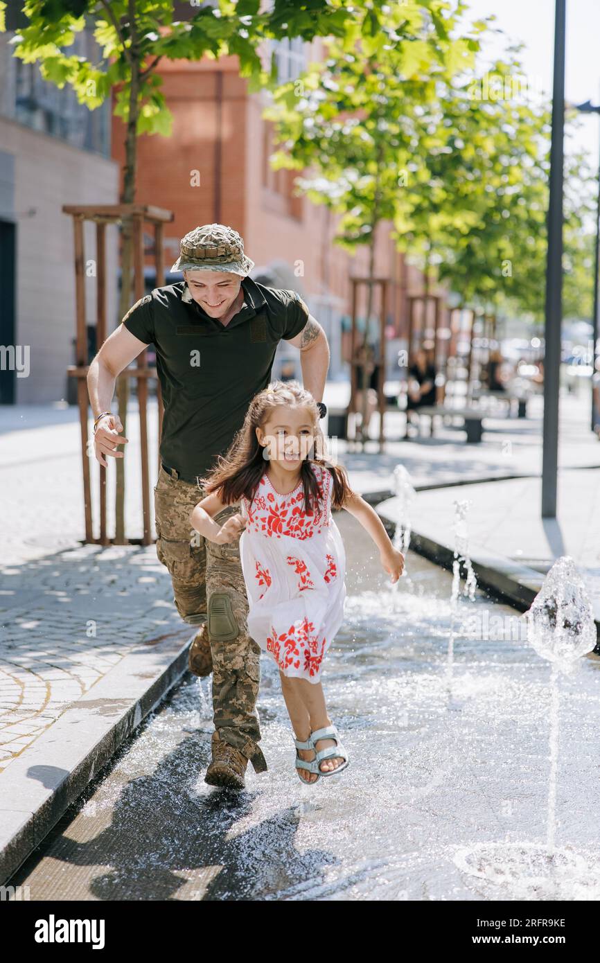 La piccola felice ragazza Ucraina incontra suo padre durante la guerra durante le sue vacanze e gioisce allegramente con lui vicino alla fontana in città. Guerra in Ucraina. Russi Foto Stock