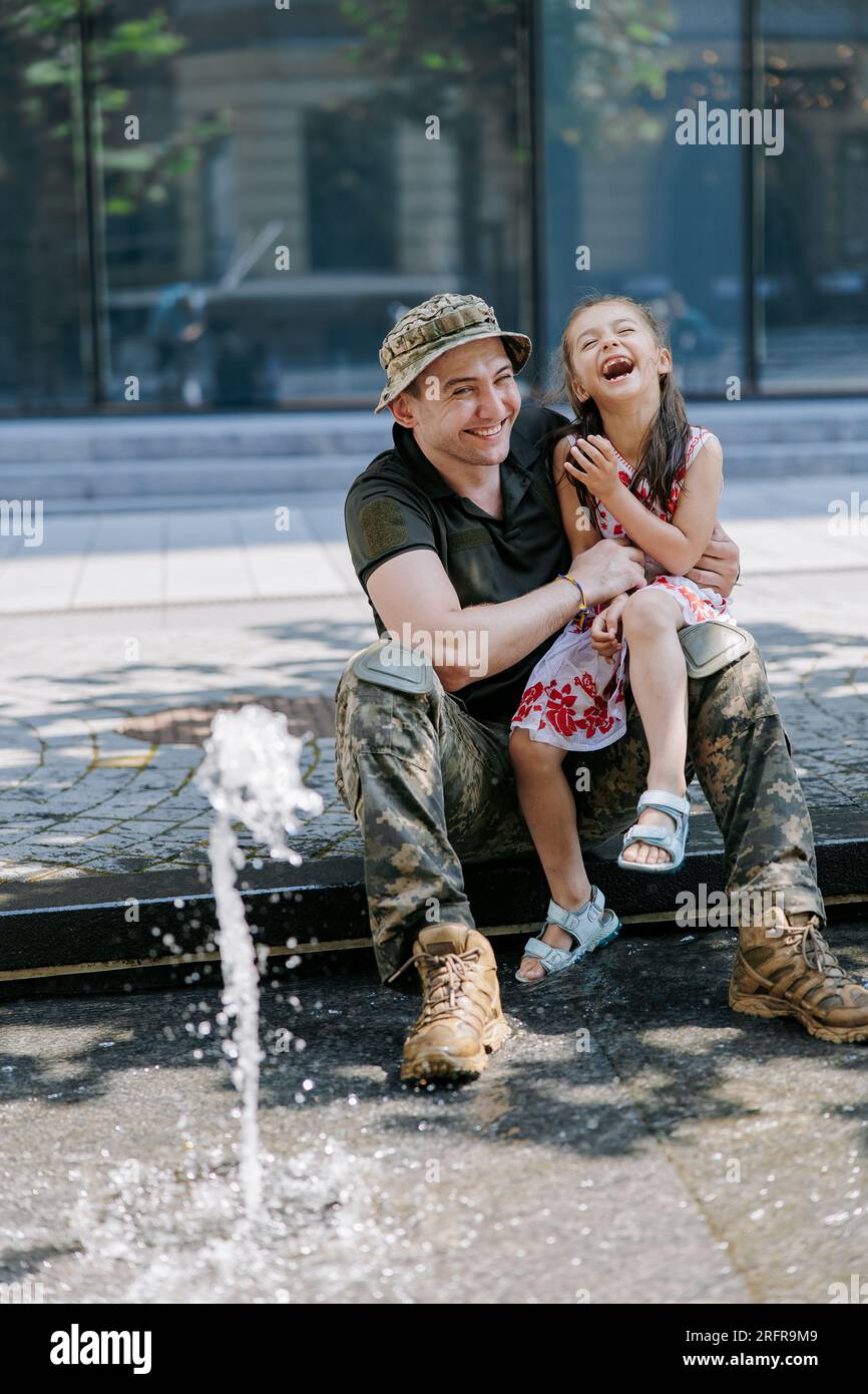 Il soldato ucraino di ritorno dalla guerra incontra la sua piccola figlia felice e allegra che gioca con lei vicino alla fontana in città. Guerra in Ucraina. Russo Foto Stock
