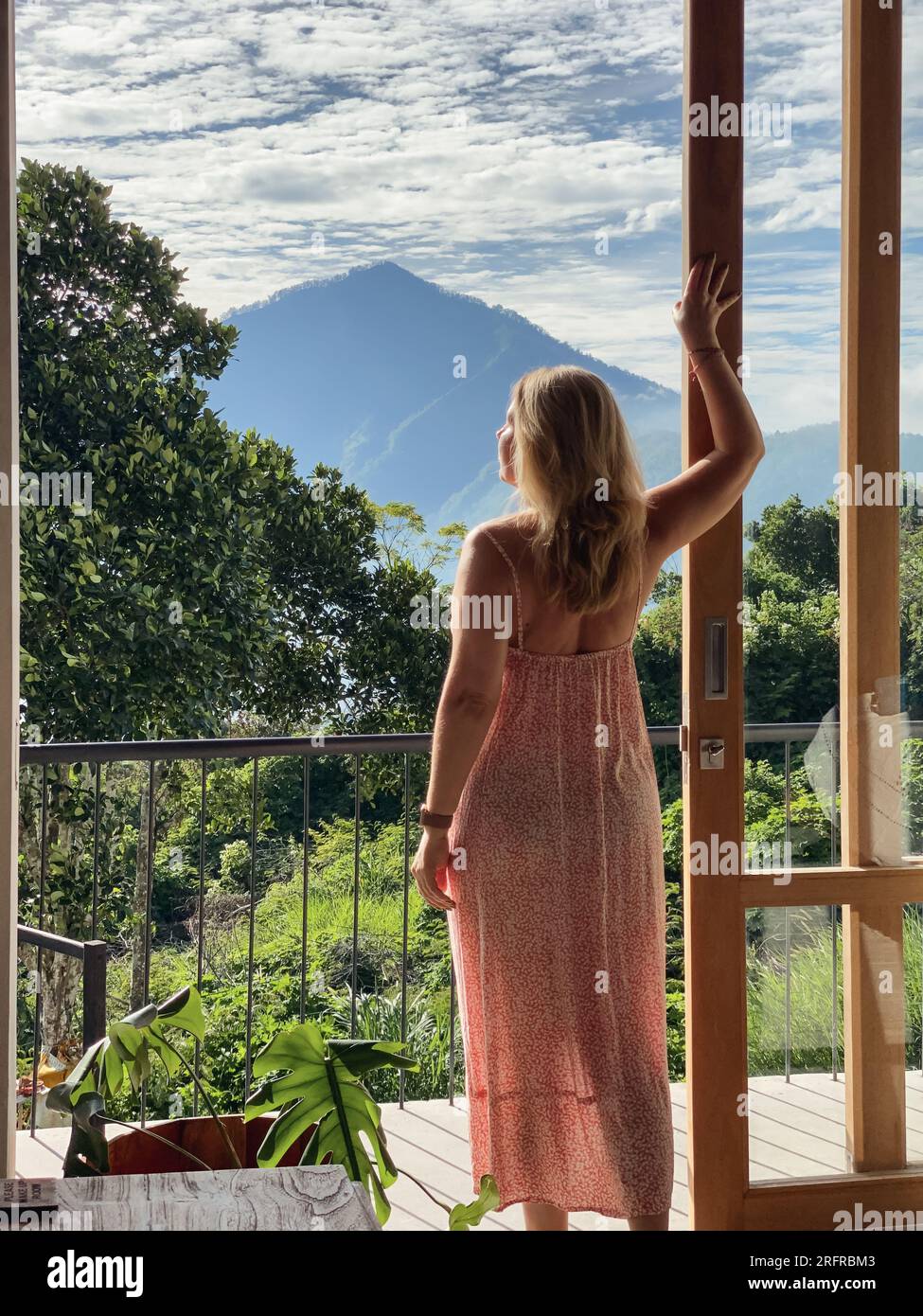 Una donna di mezza età in abito rosso si trova vicino alla porta del balcone e guarda la giungla e le montagne in una soleggiata giornata estiva Foto Stock