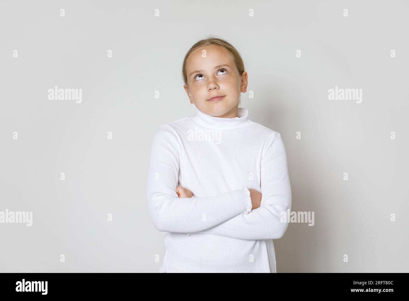 Bambino con le braccia incrociate che guarda in alto sullo sfondo della parete bianca. Giovane ragazza di 10 anni, ritratto in studio Foto Stock