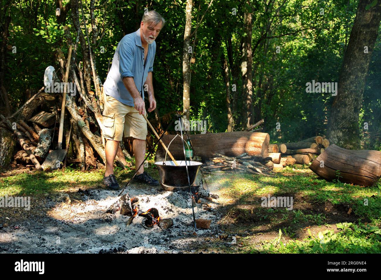 Uomo anziano che prepara cibo sul fuoco da campeggio in pentola smaltata Foto Stock