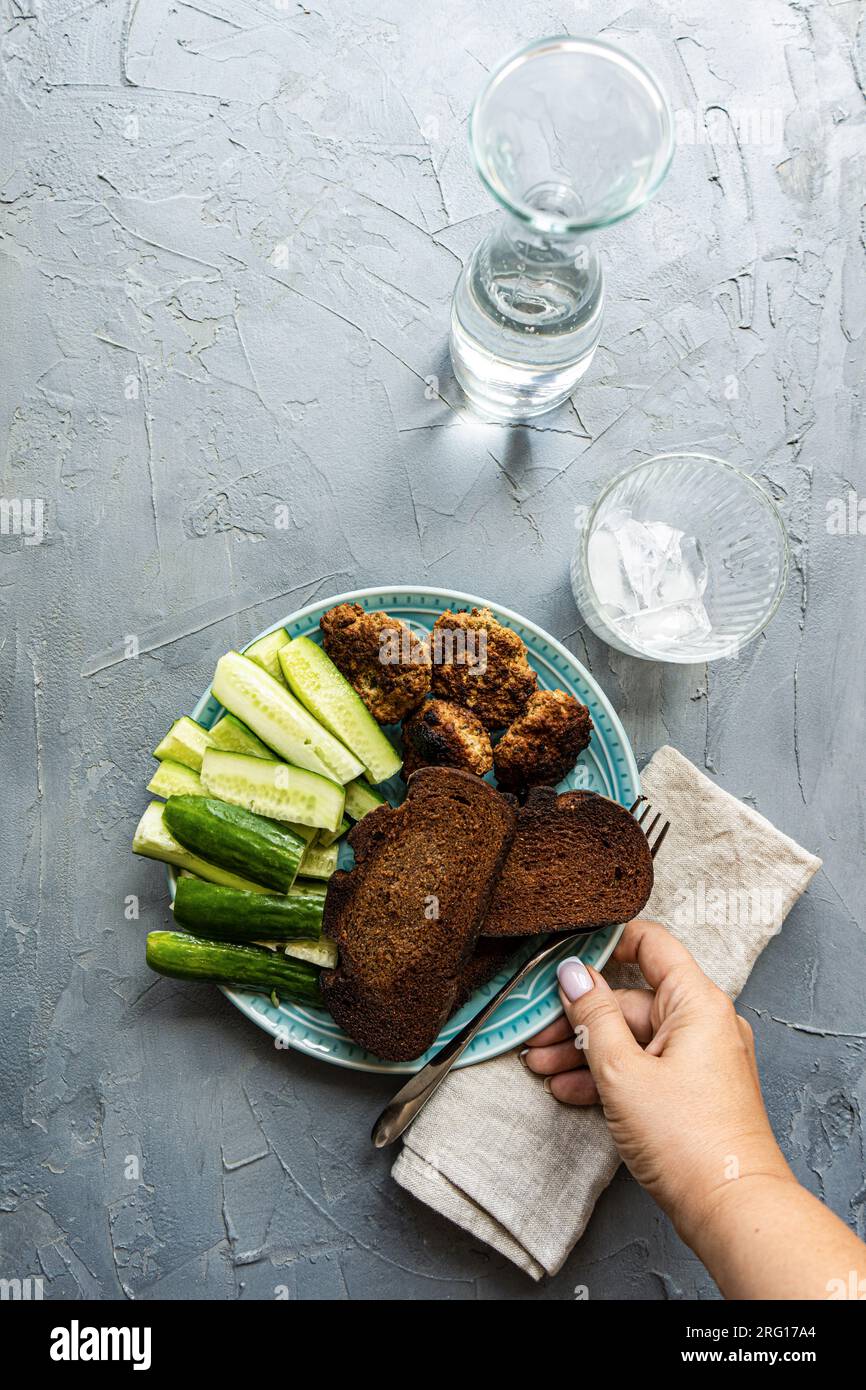 Vista dall'alto di una femmina anonima che mostra cetrioli biologici freschi, fette di segale, pane a pasta acida e cotolette di carne servite su piatto vicino al vaso d'acqua, g Foto Stock