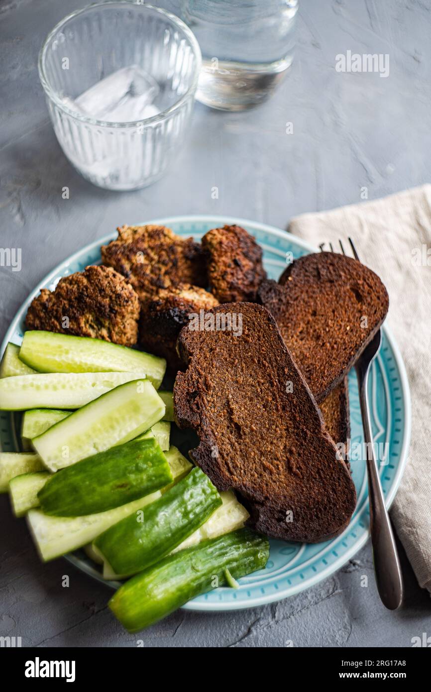 Alto angolo di cetriolo biologico fresco, fette di segale impasto acido pane e cotolette di carne servite su piatto vicino al vaso d'acqua, al vetro, alla forchetta e ai tovaglioli Foto Stock
