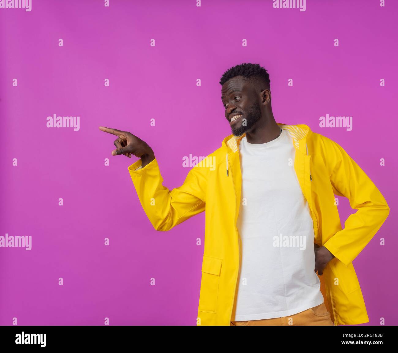 Felice giovane uomo afro-americano che guarda lontano mentre si sta in piedi con la mano in vita e punta con la mano su sfondo viola Foto Stock