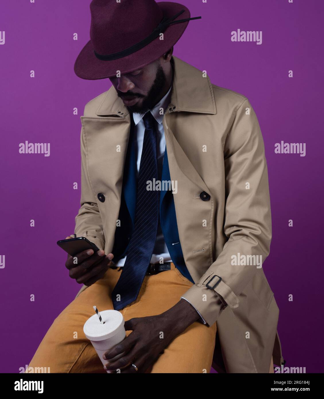 Concentrato di giovani afro-americani in elegante cravatta e cappello che guardano lo schermo del telefono cellulare mentre si siede con un bicchierino da asporto e un assegno Foto Stock