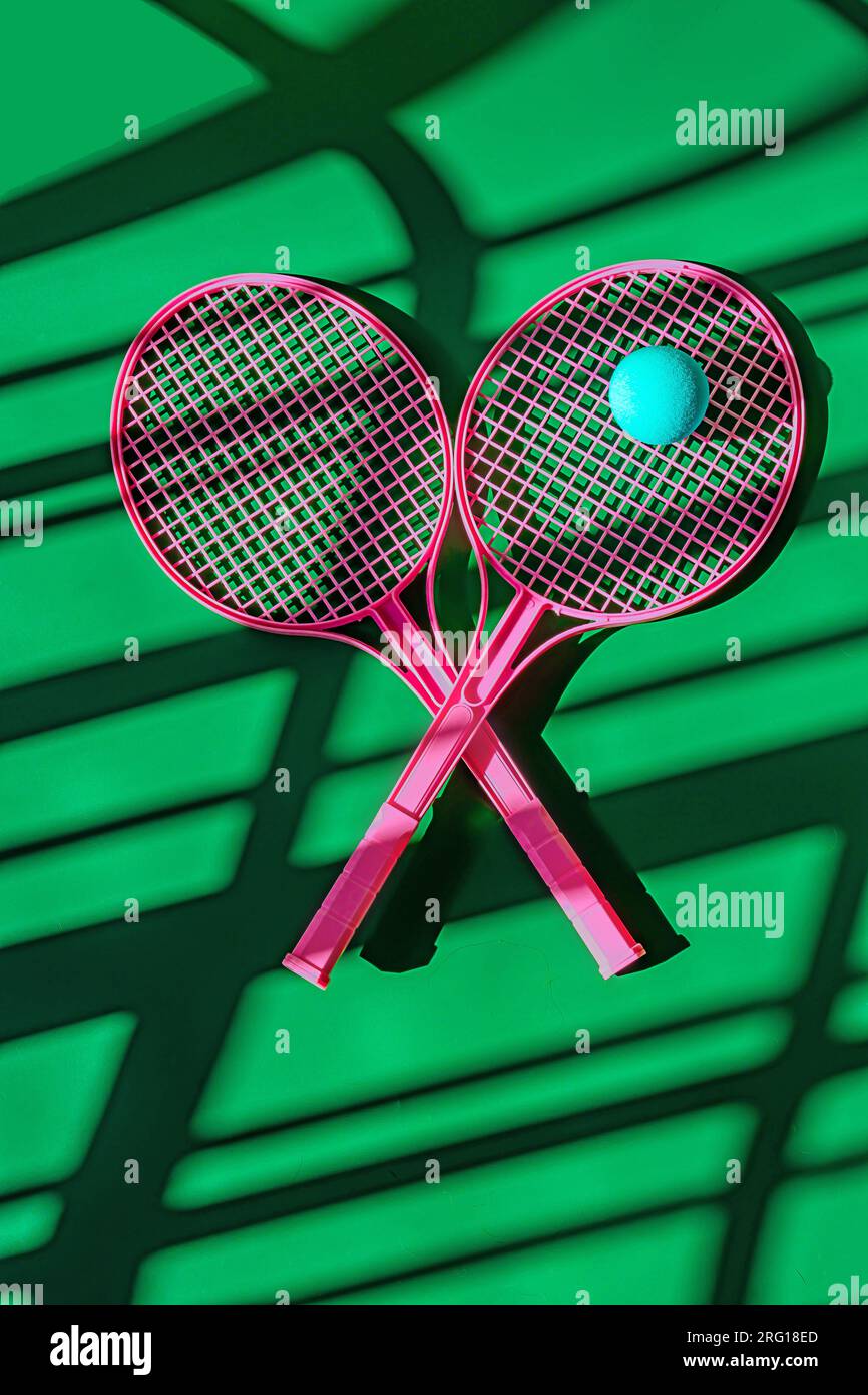 Vista dall'alto del design creativo di racchette da tennis rosa e palla blu contro il campo sportivo verde con ombre alla luce del giorno Foto Stock