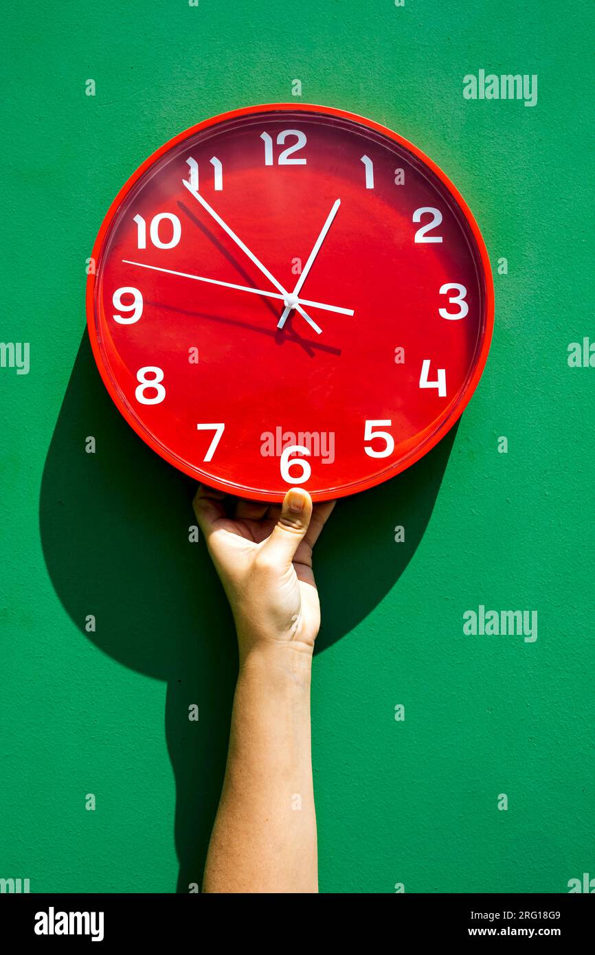 Mano di ritaglio di una persona irriconoscibile che tiene un grande orologio rosso tutto il giorno contro il muro verde Foto Stock