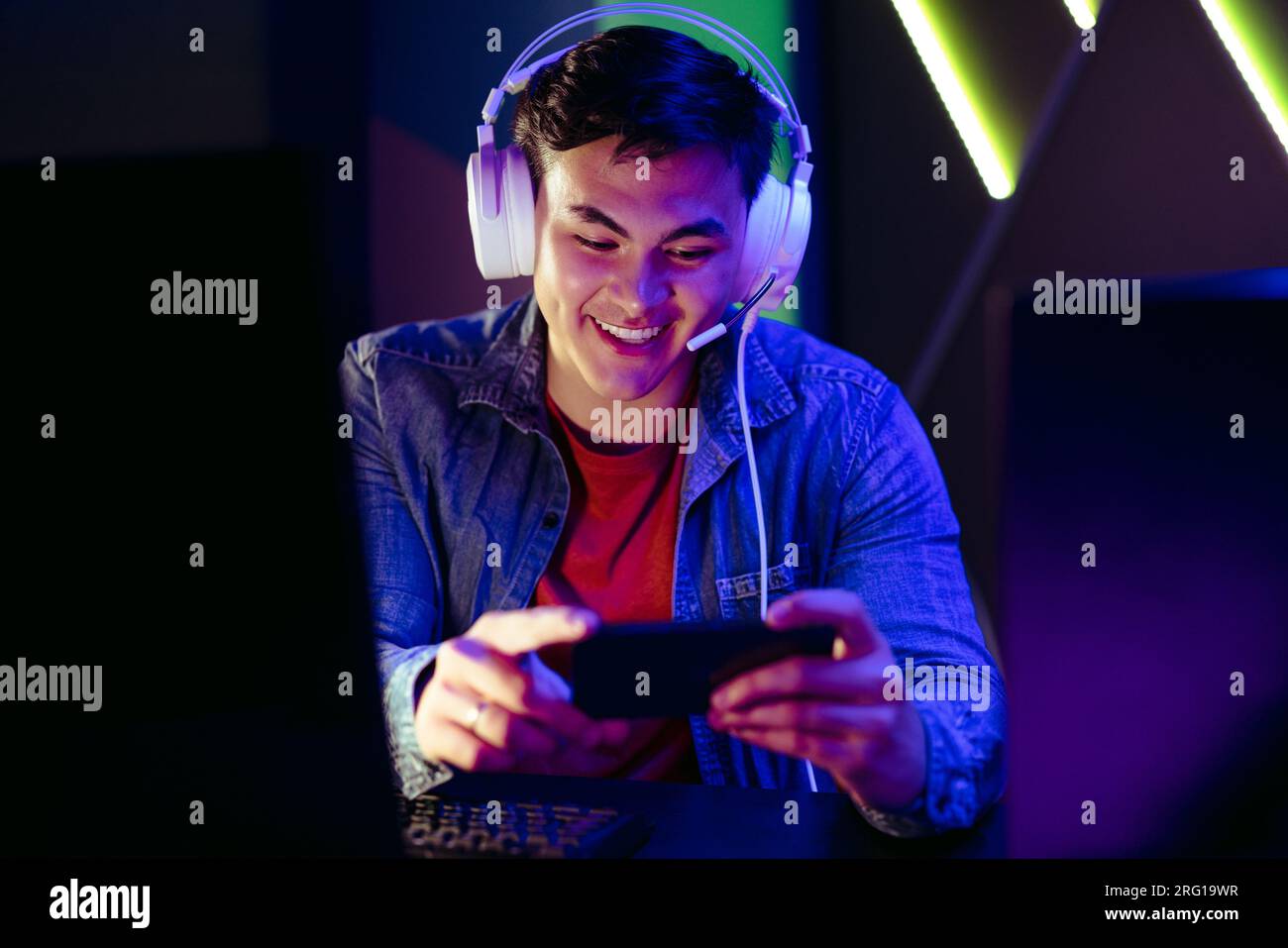 Buon giocatore di videogiochi che guarda uno streaming in diretta di un'azione di gioco sul suo smartphone. Videogamer maschile seduto alla sua stazione di gioco con le cuffie accese, che si divertono Foto Stock