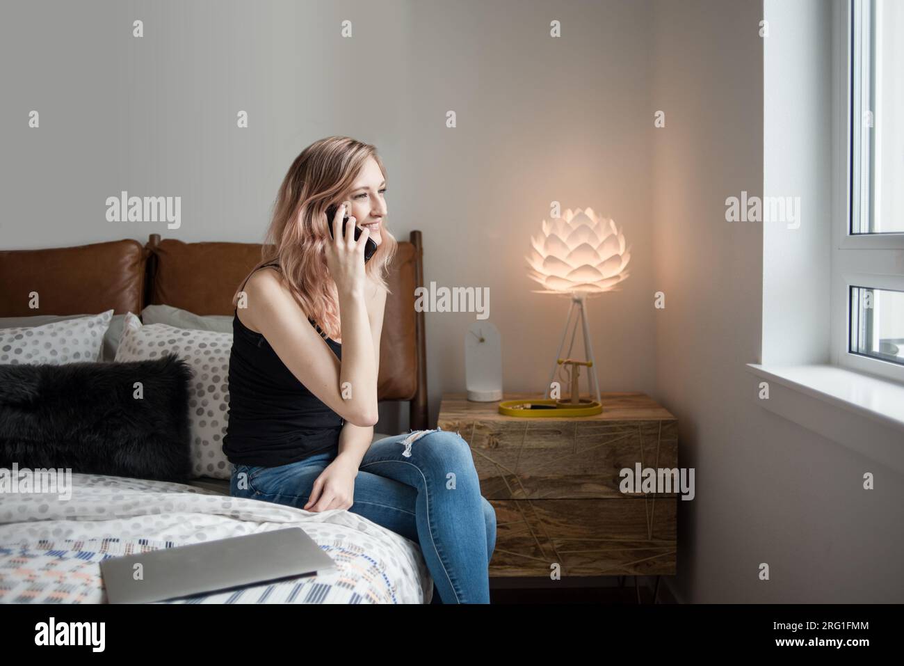 Donna che parla al telefono seduta sul lato del letto nell'appartamento Foto Stock