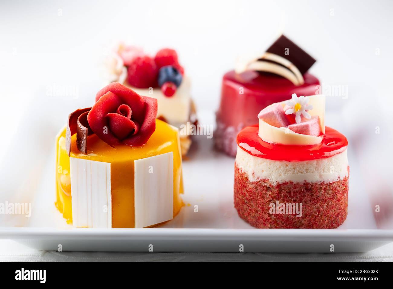 Varie mini torte su un piatto bianco. Dolci decorati con frutti di bosco freschi e fiori per le vacanze. Foto Stock