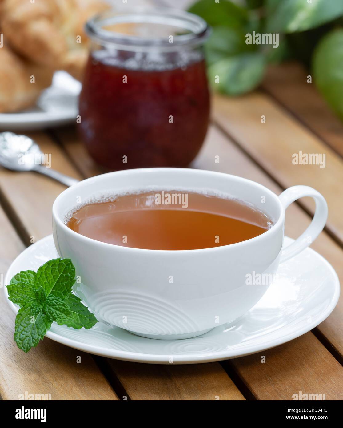 Tazza di tè con foglie di menta fresca e croissant e barattolo di marmellata sullo sfondo su un tavolo da patio in legno Foto Stock