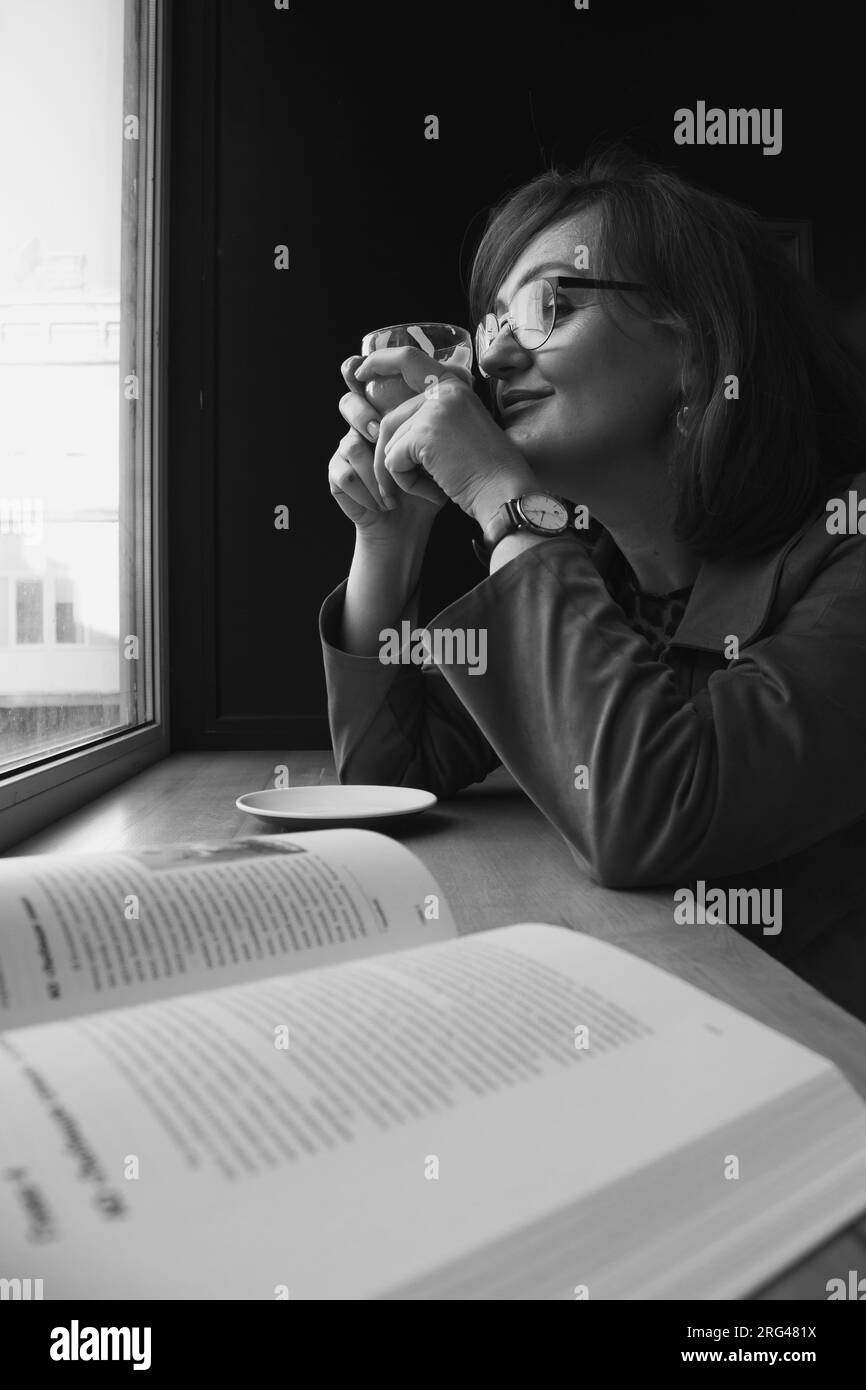 Ritratto in bianco e nero di una donna al bar. Donna in occhiali con libro aperto, bianco e nero. Donna matura che sogna in un bar. Concetto di autostima. Foto Stock