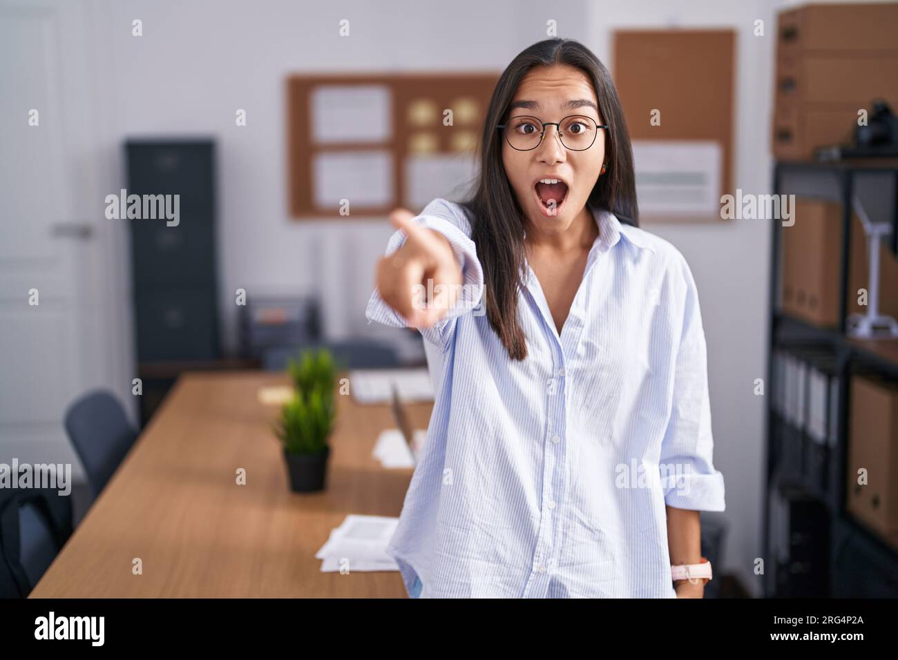 Giovane donna ispanica in ufficio che punta con il dito sorpreso davanti, bocca aperta espressione stupita, qualcosa sul davanti Foto Stock