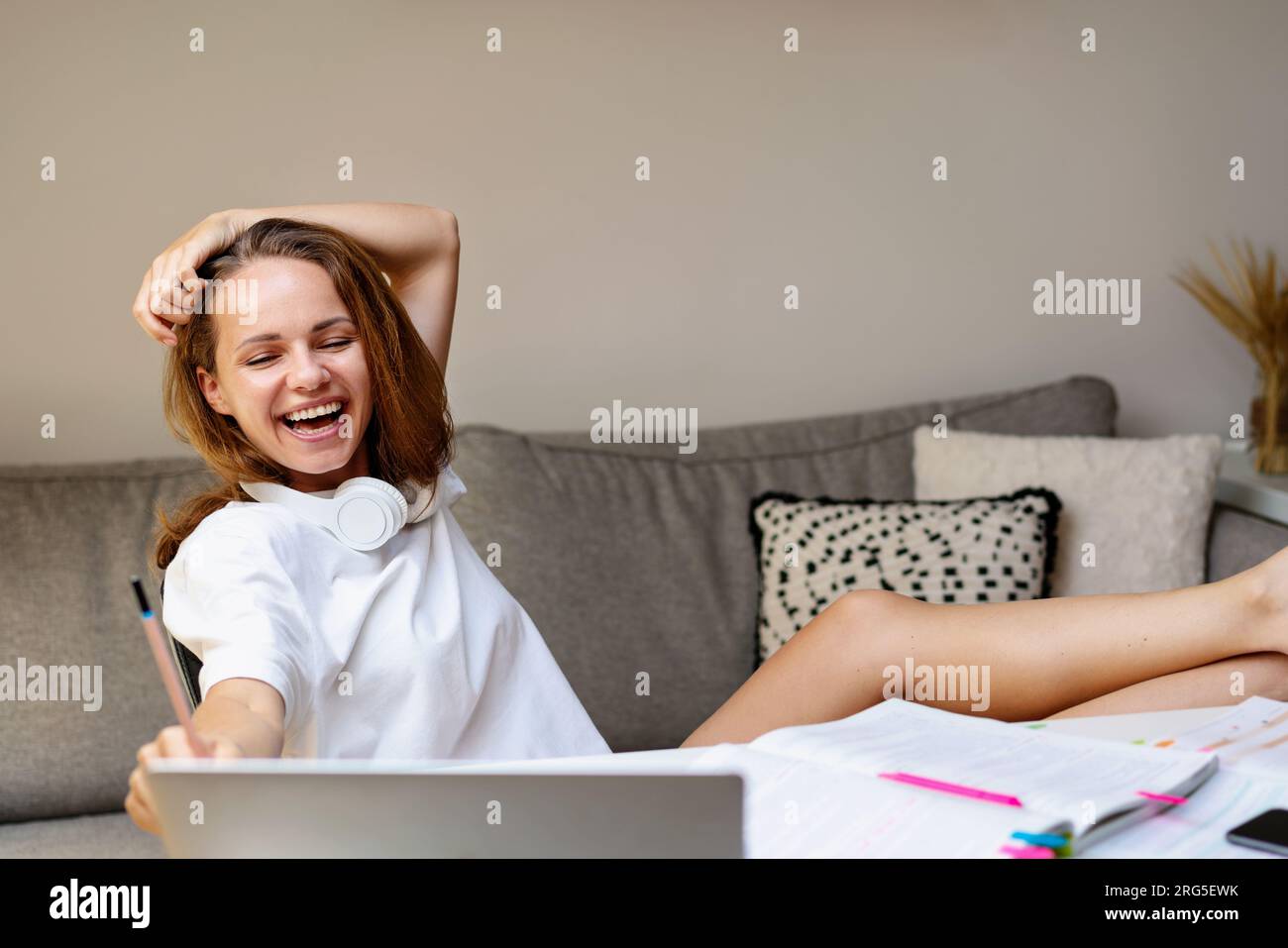 Giovane donna felice seduta al tavolo con i piedi in su a guardare video educativi online su Internet usando il suo laptop e ridendo. Foto Stock