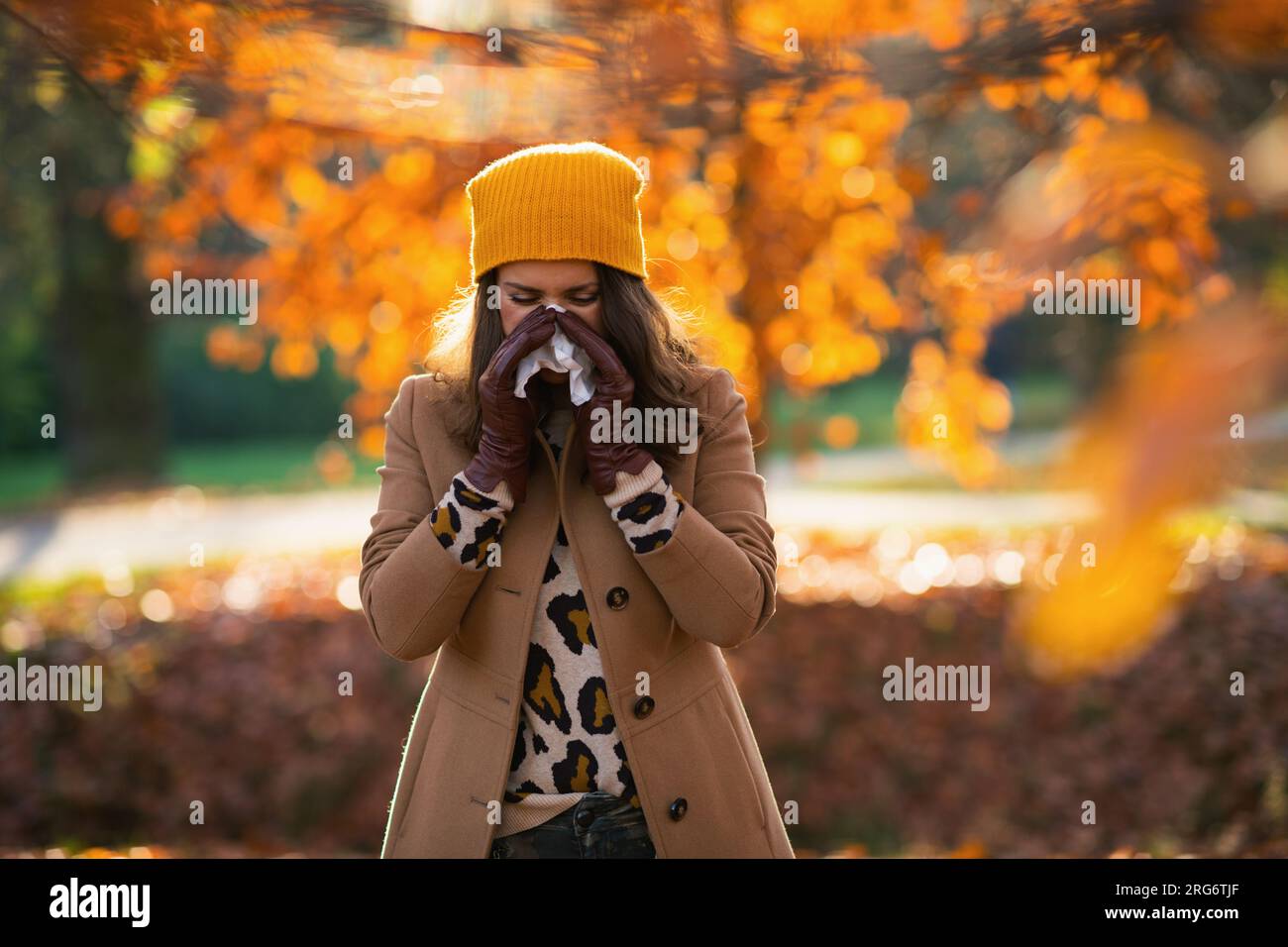 Ciao, september. elegante donna di mezza età con cappotto beige e cappello arancione con tovagliolo che soffia il naso all'esterno nel parco cittadino in autunno. Foto Stock