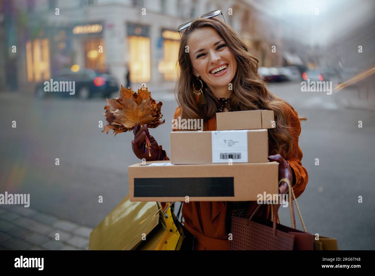 Ciao november. felice donna moderna di 40 anni in trench marrone con pacchi, borse della spesa e foglie gialle autunnali in città. Foto Stock