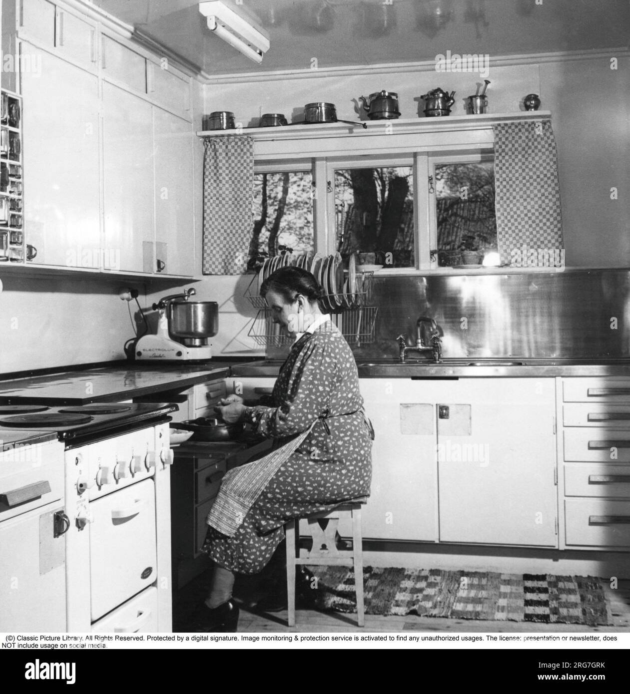 Negli anni '1940 Una donna anziana in una tipica cucina degli anni '1940 con i dettagli dell'interno e gli oggetti che sono molto rappresentativi del decennio. Un assistente di casa, una macchina da cucina, è un apparecchio elettrico la cui funzione di base è di mescolare e lavorare l'impasto ed è visto in piedi sul banco. In realtà, l'assistente domestico è una parola di marca presa dal modello Electrolux chiamato Assistant. Electrolux ha registrato la parola Assistant come marchio. A differenza, ad esempio, dei robot da cucina e delle frustate elettriche, la lavorazione in un assistente avviene a una velocità significativamente inferiore, il che Foto Stock