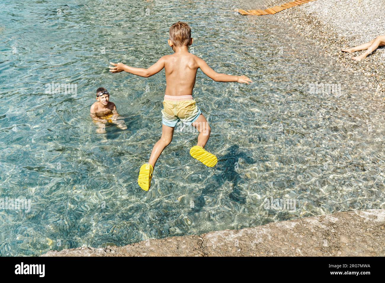 Il ragazzino salta volando dal molo in una posizione divertente nell'acqua di mare. Il bambino in età prescolare ama trascorrere le vacanze con la famiglia al mare Foto Stock