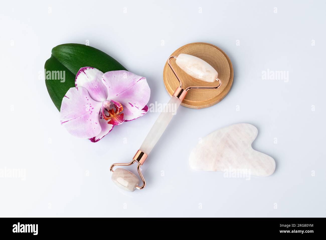 Massaggiatore Gua Sha in pietra naturale. Rullo facciale al quarzo, fiore di orchidea rosa, foglia verde, sfondo bianco, vista dall'alto. Strumento di massaggio del viso. Cura della pelle, S Foto Stock