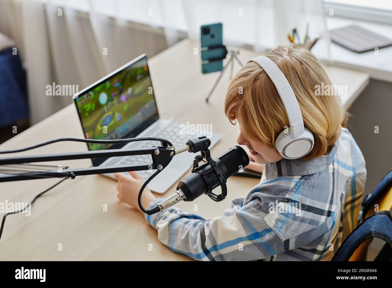 Ragazzino biondo con cuffie che parla nel microfono mentre si siede davanti al computer portatile con un videogioco sullo schermo e si gioca Foto Stock
