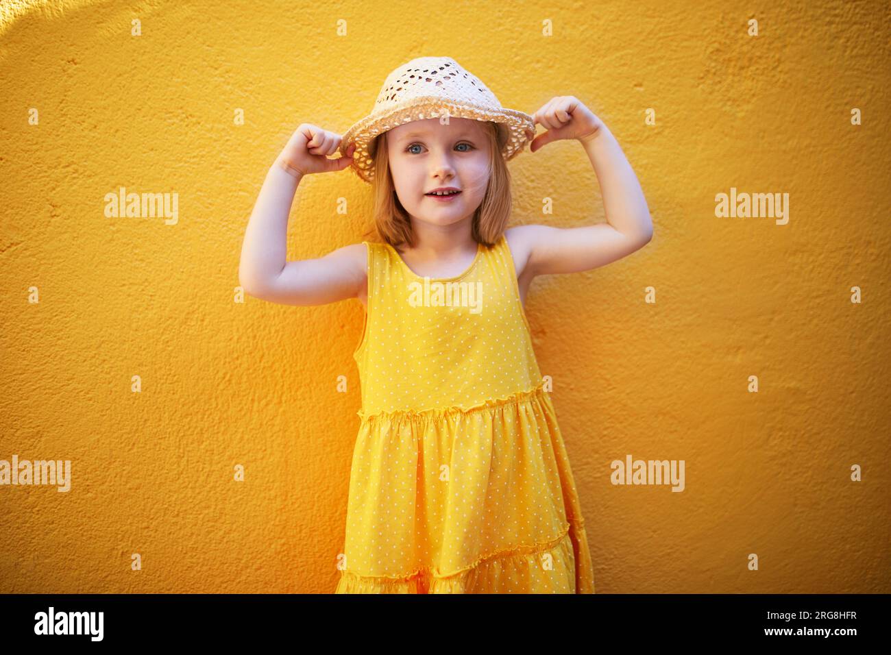 Allegra bambina di tre anni con vestito giallo e cappello di paglia che posa contro il muro giallo. Bambino felice in un giorno d'estate. Attività estive o vacanze Foto Stock