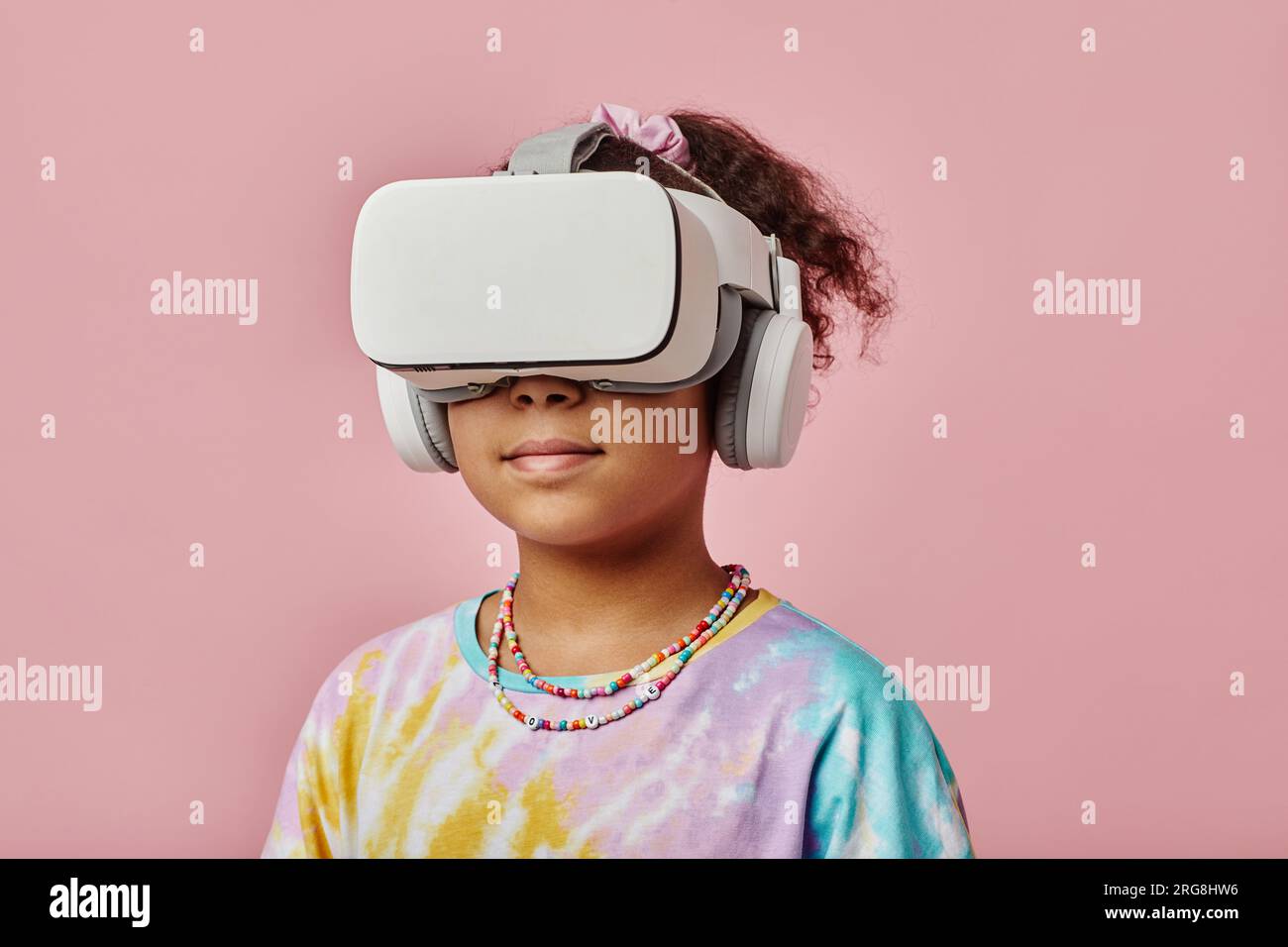Studentessa con visore VR in piedi di fronte alla fotocamera su sfondo rosa durante il videogioco mentre posa isolata Foto Stock