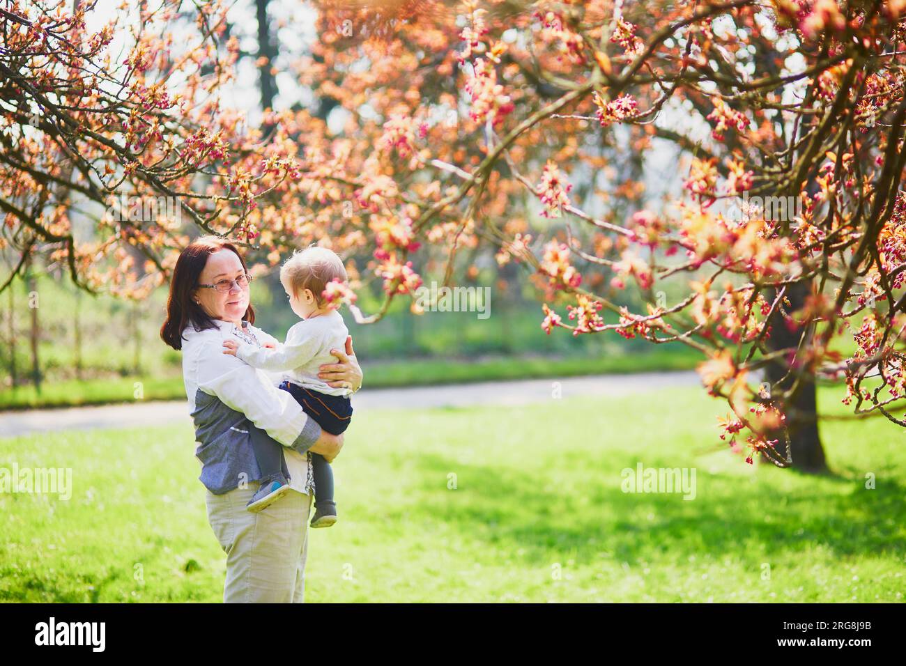 Donna di mezza età che tiene una bambina di 1 anno in parco in un giorno di primavera. La nonna e la nipote si godono la stagione della fioritura dei ciliegi. Attività di famiglia Foto Stock