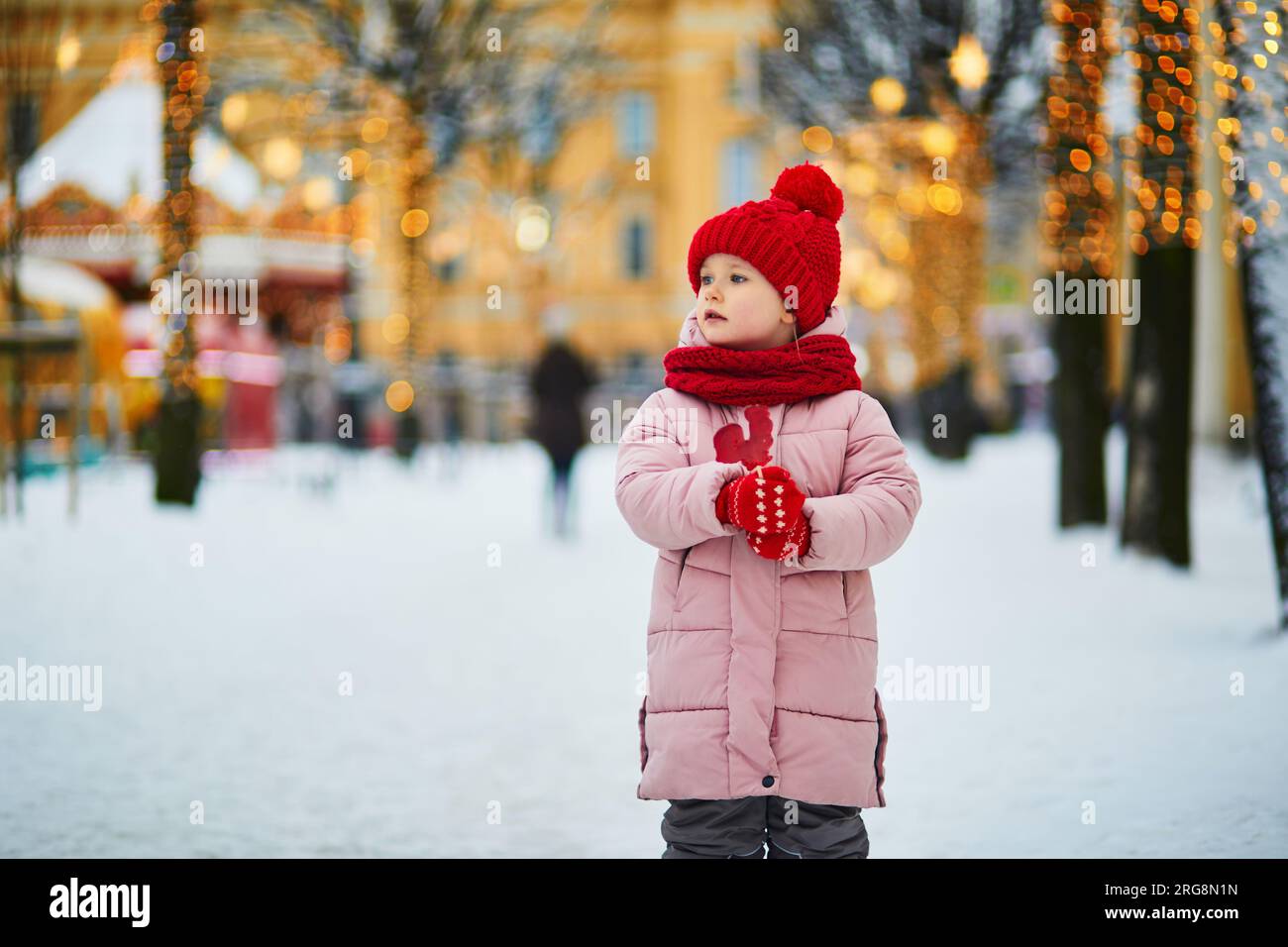 Allegra e allegra bambina in età prescolare in rosso snood e cappello che si diverte al mercatino di Natale e mangia lecca-lecca a forma di gallo Foto Stock