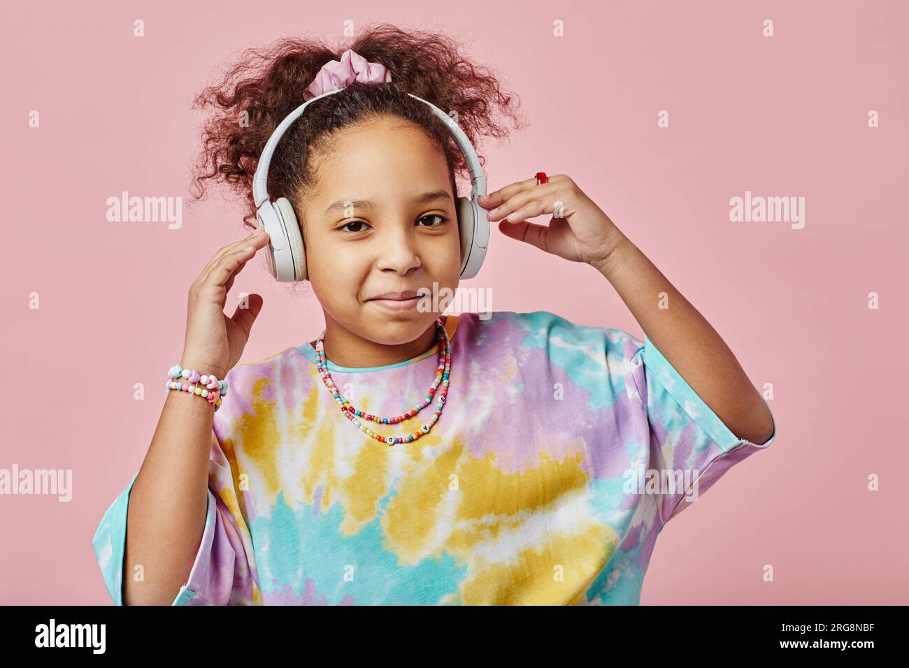 Simpatica studentessa che indossa le cuffie da gioco e ascolta musica mentre si trova davanti alla fotocamera su sfondo rosa in studio Foto Stock