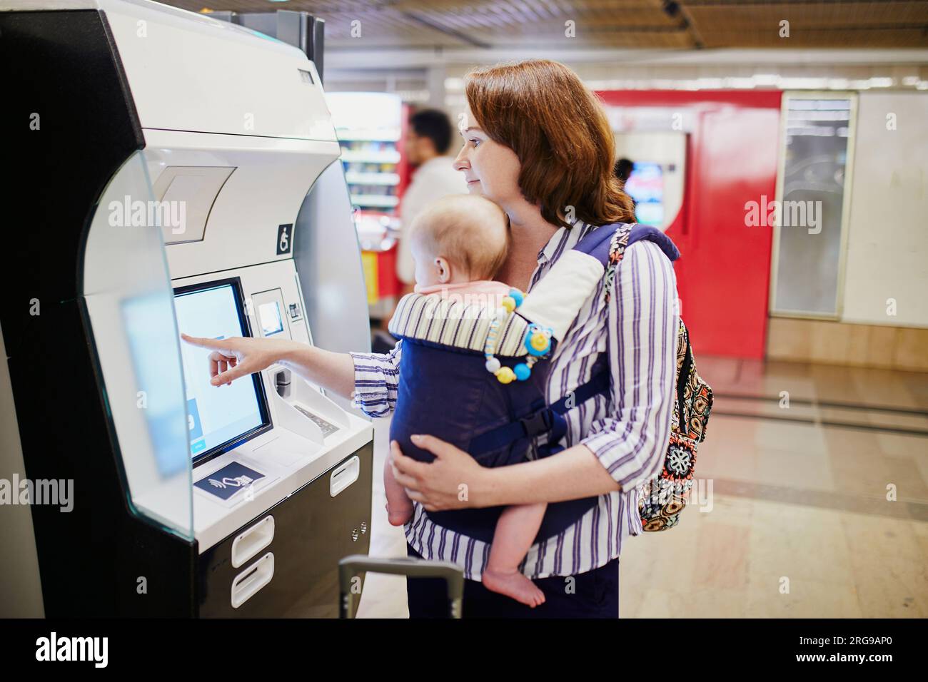 Una giovane madre con il suo bambino che prende i biglietti alla stazione ferroviaria. Una donna con bambino piccolo nel portabicchieri che effettua il check-in in aeroporto. TR Foto Stock