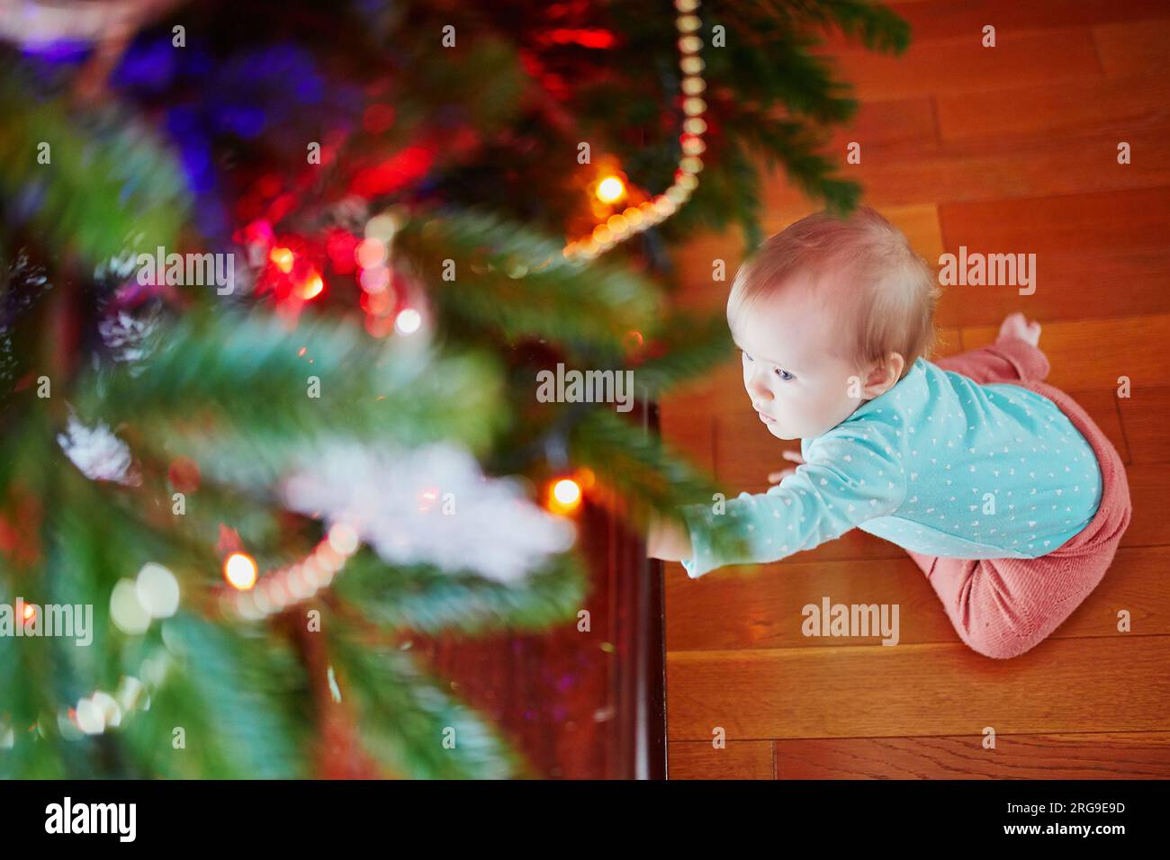 Bambina seduta per terra sotto l'albero di Natale. Bambino che strizza a casa ed esplora le cose intorno a lei. Vacanze con bambini piccoli Foto Stock