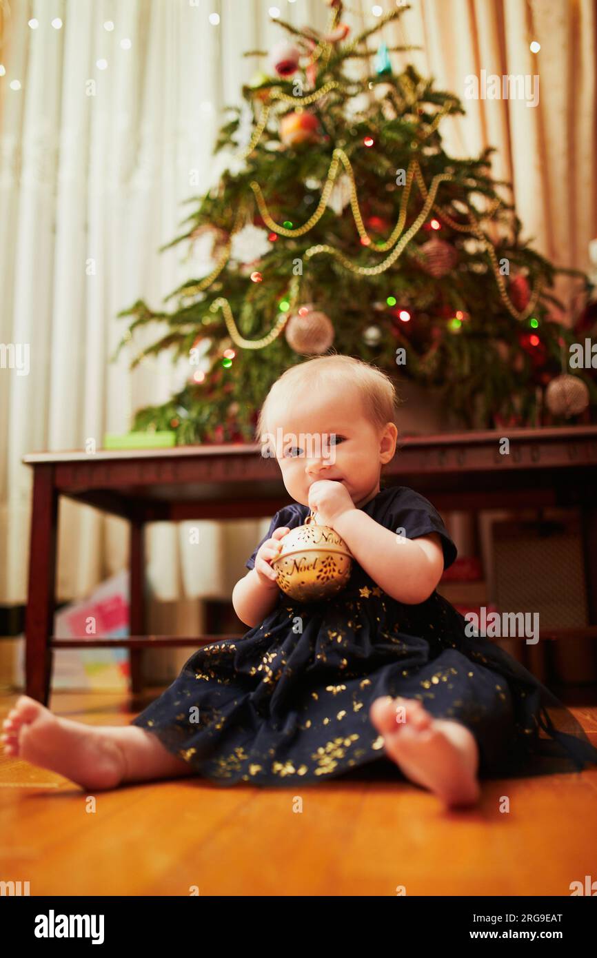 Bambina seduta in un bel vestito sul pavimento sotto l'albero di Natale. Bambino che strizza a casa ed esplora le cose intorno a lei. Vacanze con l Foto Stock