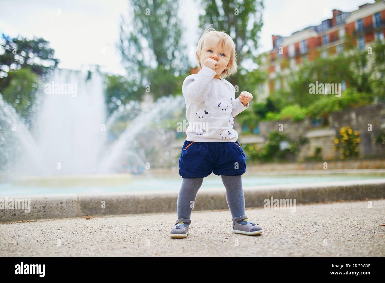 Adorabile bambina all'aperto che mangia pane nel parco. Bambino che gioca vicino alla fontana in una strada di Parigi. Attività all'aperto per bambini Foto Stock