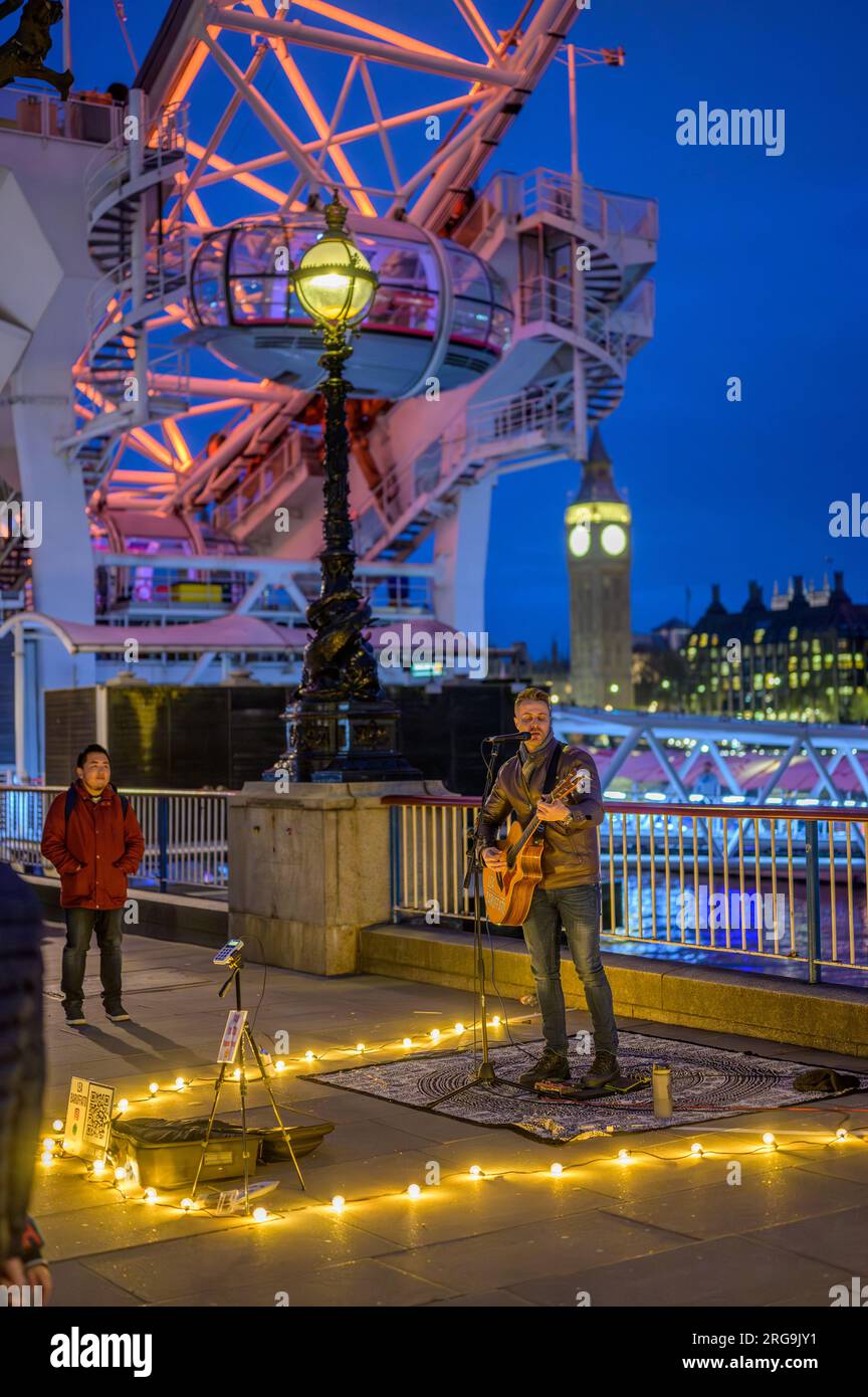 LONDRA - 22 aprile 2023: Notte incantevole: Busker intrattiene i turisti dal London Eye a Southbank, luci calde contro il cielo blu profondo, con il Big Ben Foto Stock