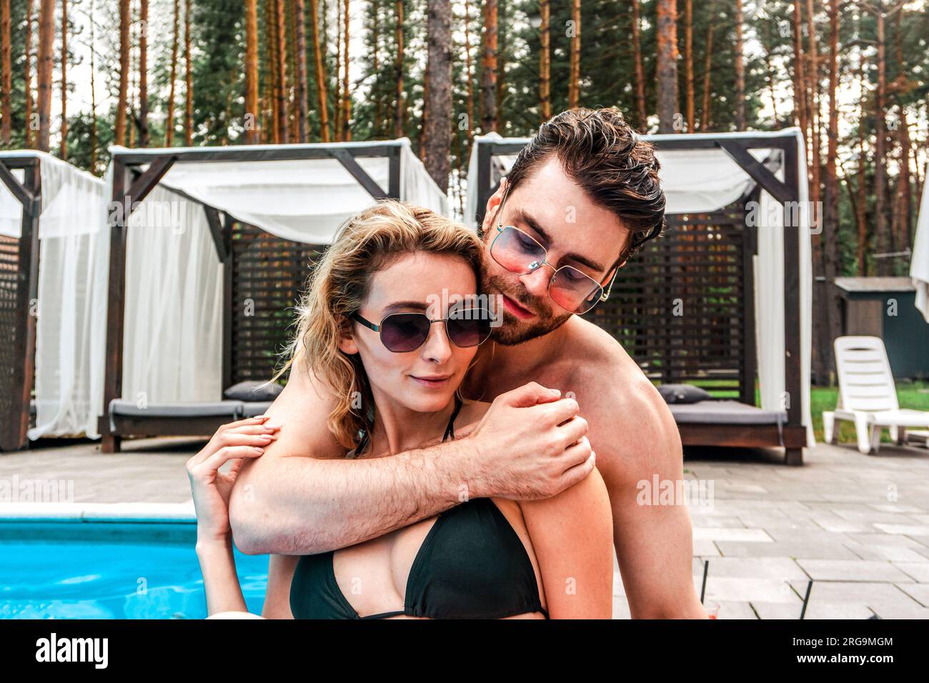 Uomo innamorato che abbraccia la sua ragazza con gli occhiali da sole mentre si siede dietro di lei a bordo piscina. Romanticismo e vacanze estive Foto Stock