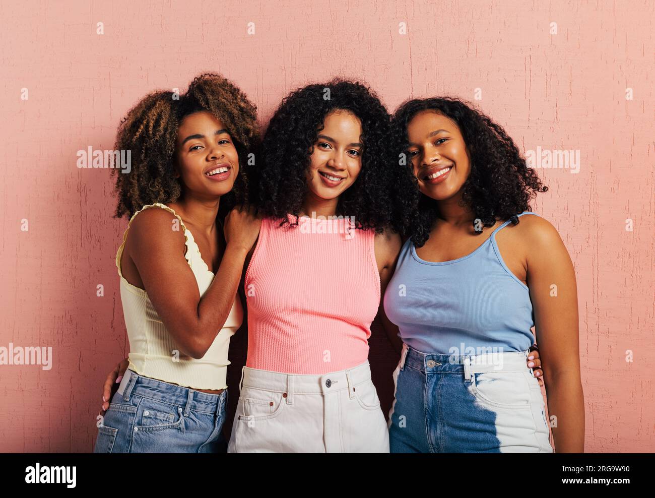 Tre bellissime donne sorridenti con capelli ricci in piedi insieme su una parete rosa Foto Stock