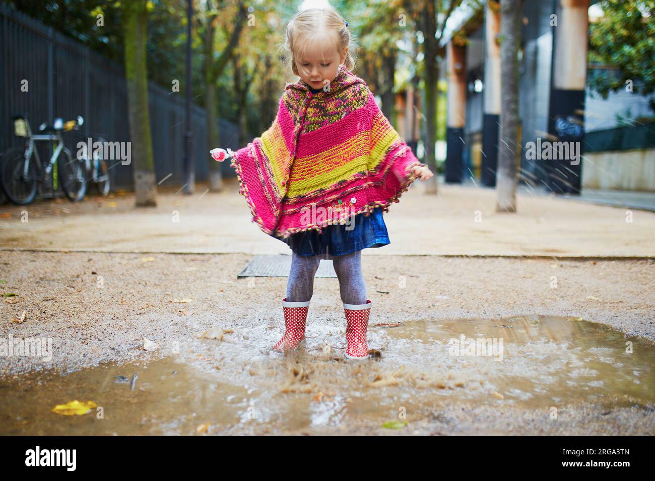 Bambino che indossa stivali da pioggia rossi e salta in pozzanghera in autunno. Adorabile bimba che si diverte con acqua e fango nel parco in una giornata di pioggia. Esterno Foto Stock