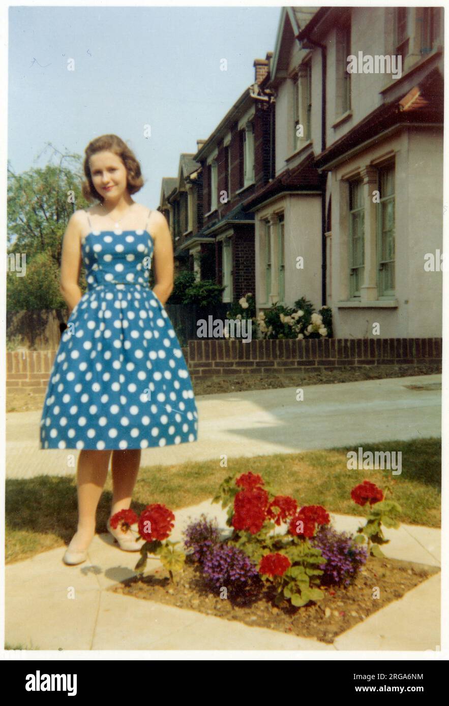 Una giovane signora in un abito blu e mentre pois polka in piedi in un pulitissimo giardino suburbano britannico anteriore - accanto a un piccolo a forma di diamante fiorito contenente alcuni garofani rossi - settembre, 1959 Foto Stock