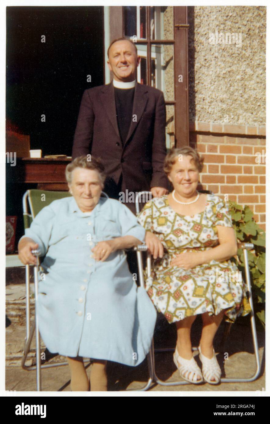 Due donne più anziane che siedono appena fuori della porta posteriore di una casa britannica suburbana in sedie pieghevoli leggere sono unite dal vicario locale per una fotografia di gruppo - ottobre, 1959 Foto Stock