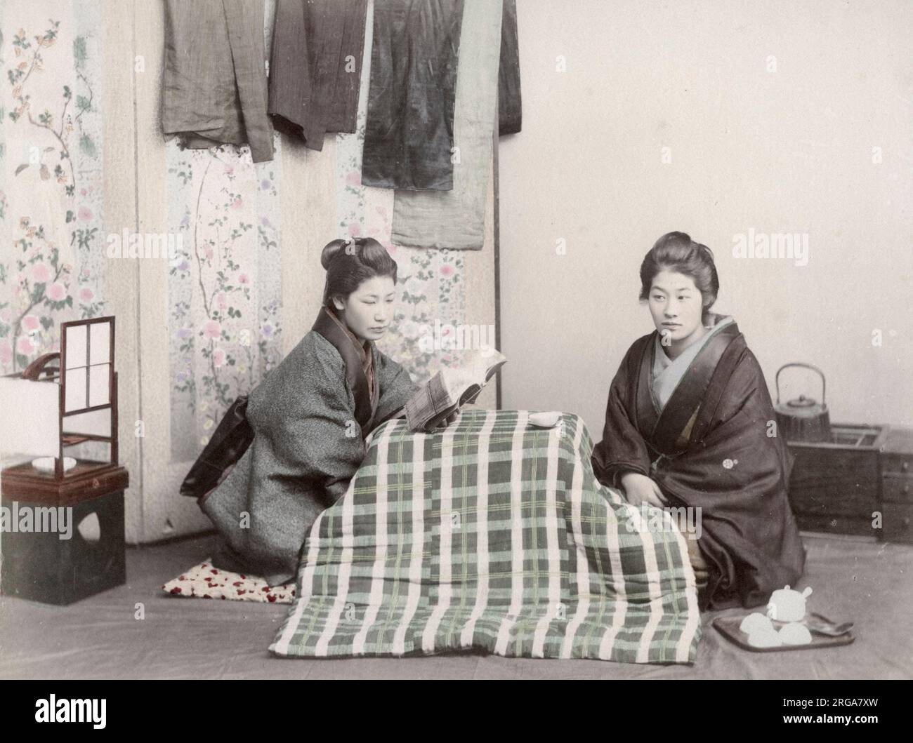 Due giovani donne che leggono e rimangono calde sotto le coperte intorno ad una stufa sotto un tavolo di kotatsu, Giappone. Vintage 19th ° secolo fotografia. Foto Stock