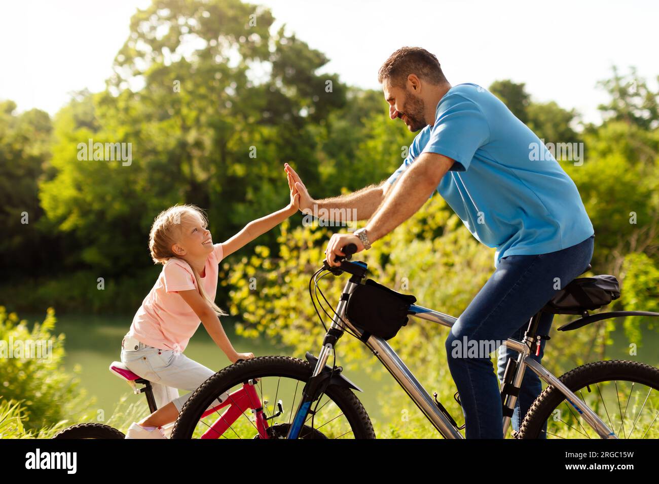 Papà e sua figlia danno cinque premi mentre si pedala nel soleggiato parco, goditi il tempo libero insieme, con vista laterale Foto Stock