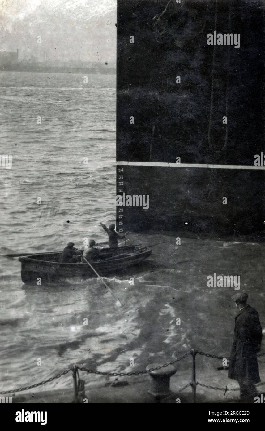 Tre uomini in una piccola barca a remi svaniti dalla massiccia prua di una grande nave a vapore presso (probabilmente) Liverpool Docks, Merseyside. I numeri chiaramente visibili disposti in una linea verticale sono segni di sformo, che misurano la distanza tra il fondo dello scafo (la chiglia) e la linea di galleggiamento. Foto Stock