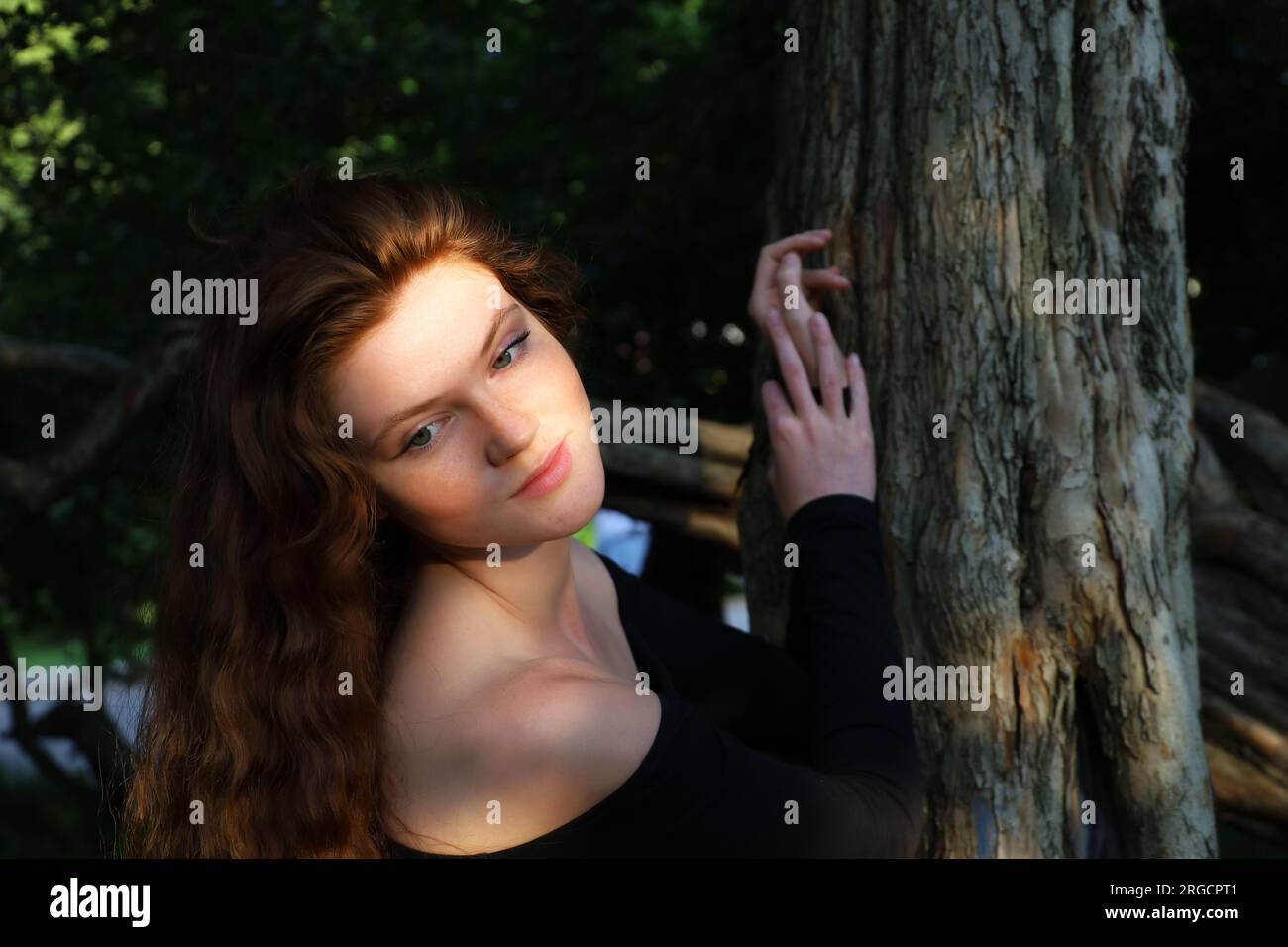 Ritratto di una sensuale ragazza allo zenzero con lunghi capelli rossi e lentiggini in piedi vicino all'albero alla luce del sole serale. Giovane modello seducente Foto Stock