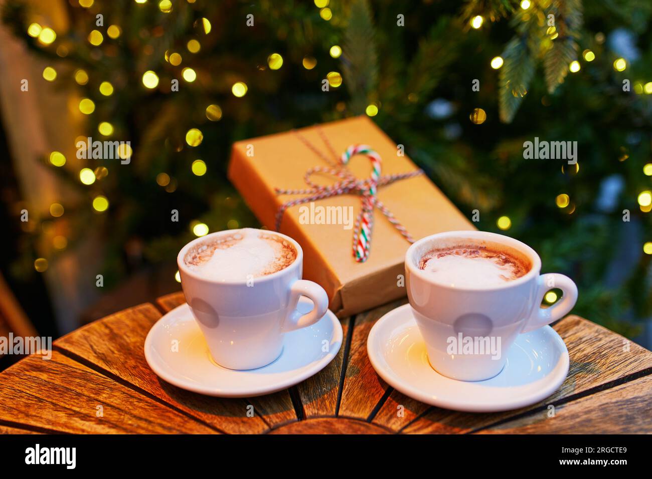 Due tazze di caffè e un regalo di Natale avvolto su un tavolo di legno del caffè o del ristorante vicino all'albero di Natale decorato con ghirlande chiare Foto Stock