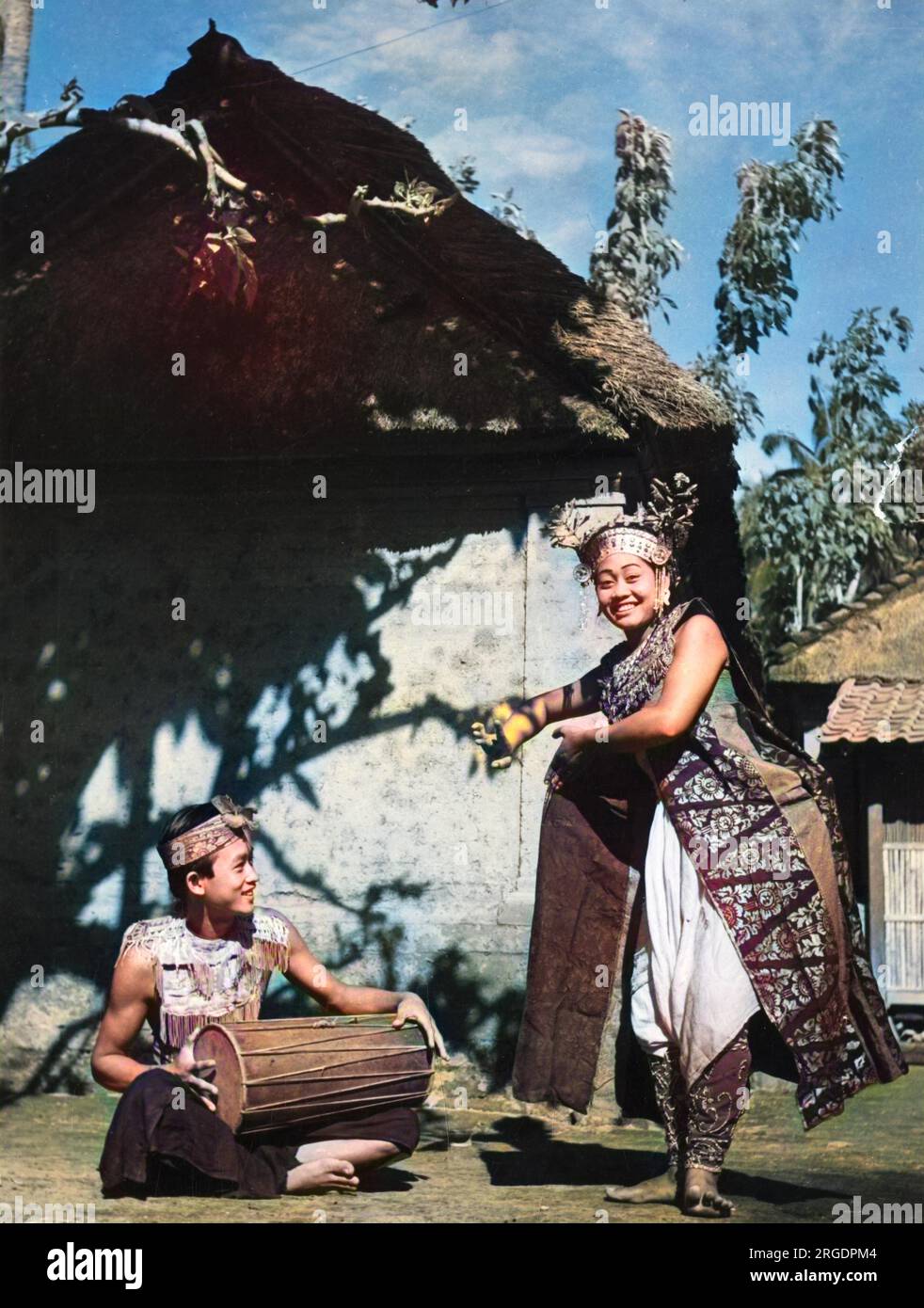 Bali, Indonesia: Una ballerina inizia la sua routine come giullare di corte, rivestita di sete riccamente colorate e decorate. Un ragazzo batterista siede accanto a lei. Foto Stock