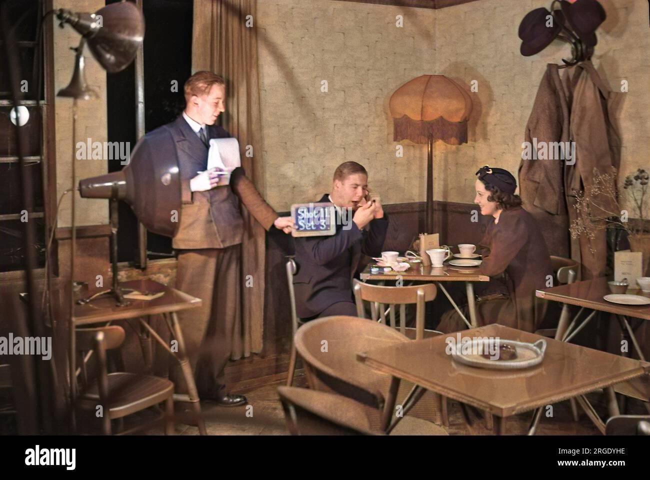 Cast e la troupe su un set cinematografico -- un uomo e una donna seduti a un tavolo, mentre un altro uomo tiene una ardesia (Shot 11, set 3). Foto Stock