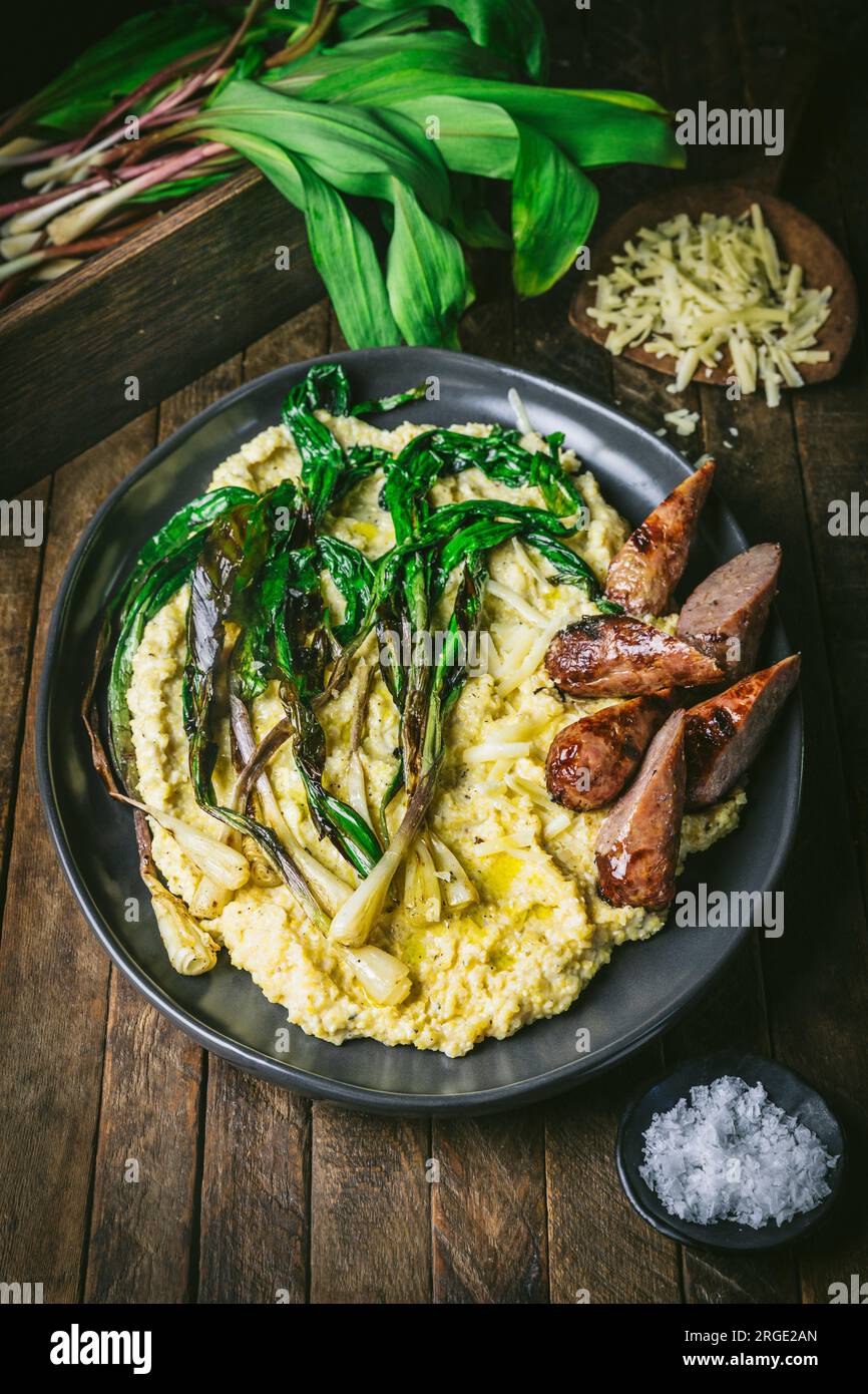Polenta, rampe grigliate e salsiccia a fette su un piatto con rampe fresche e formaggio grattugiato sullo sfondo Foto Stock