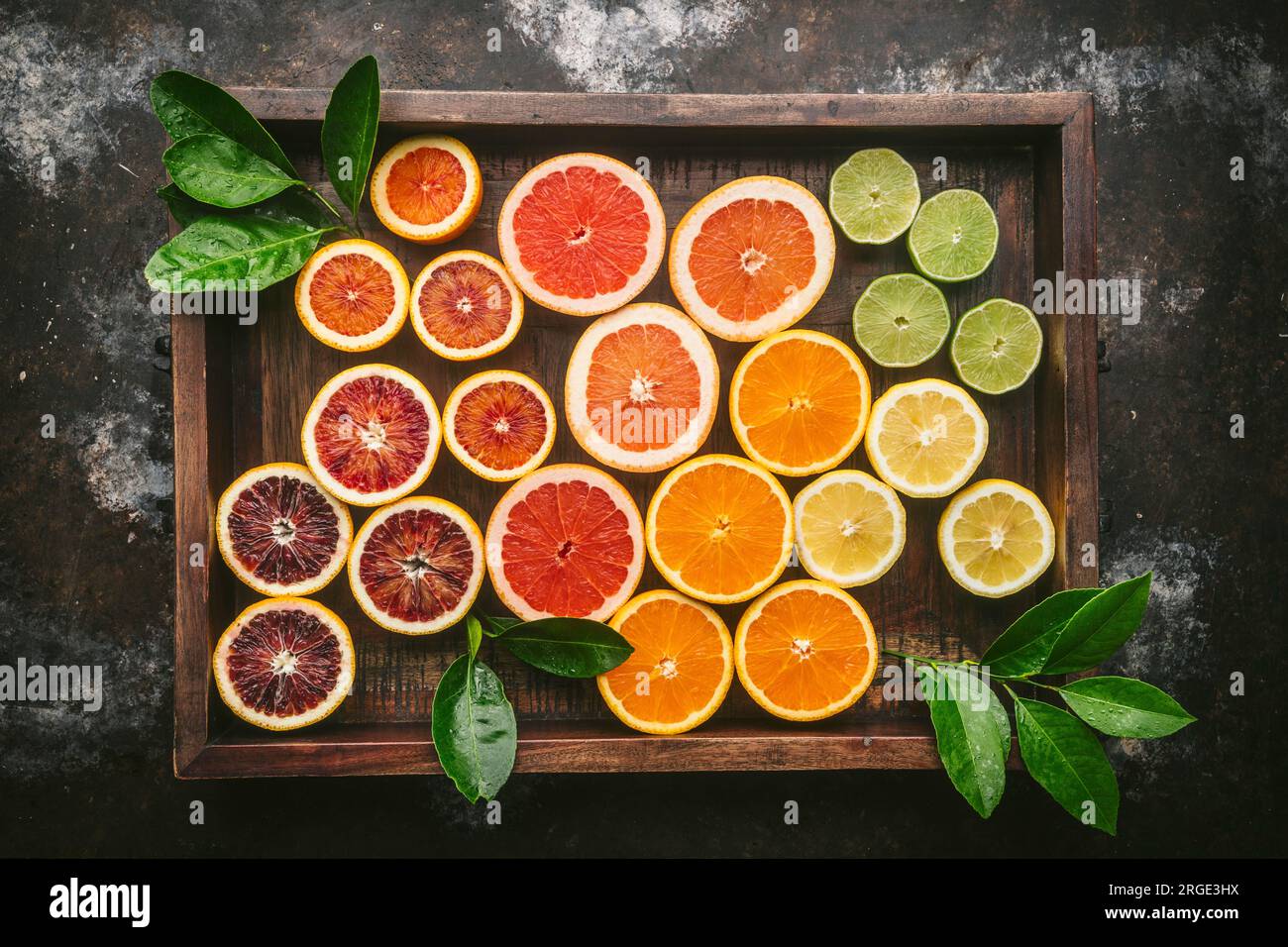 Agrumi (arancia, arancia rossa, pompelmo, limone, lime) tagliati a metà e disposti per colore in scatola di legno rustico con foglie di agrumi fresche Foto Stock