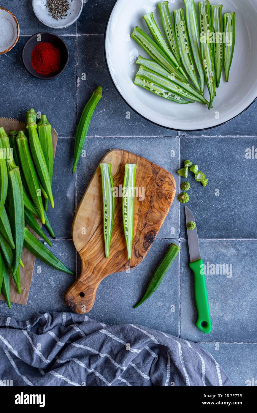 Fette di okra a forma di patatine fritte in una ciotola bianca e una cialda di okra tagliata a metà longitudinalmente su un tagliere, un coltello sul lato. Foto Stock