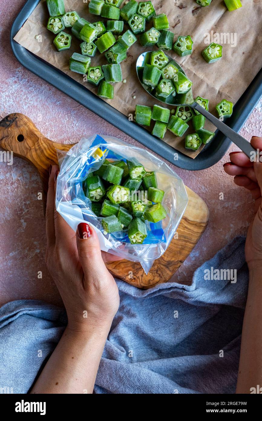 Le mani trasferiscono le fette di okra fresche da una teglia da forno rivestita di carta da forno in un sacchetto per congelatore. Foto Stock